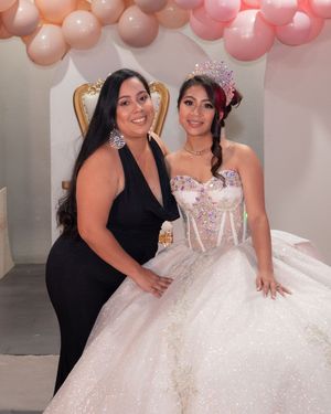 Two women pose together at quinceañera, one in black dress and one in sparkling white ball gown with floral crown.