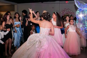 Quinceañera girl in white ball gown dances with young girls in pink dresses on decorated dance floor.