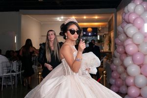 Quinceañera girl in white ball gown and tiara poses confidently wearing sunglasses near balloon column entrance.
