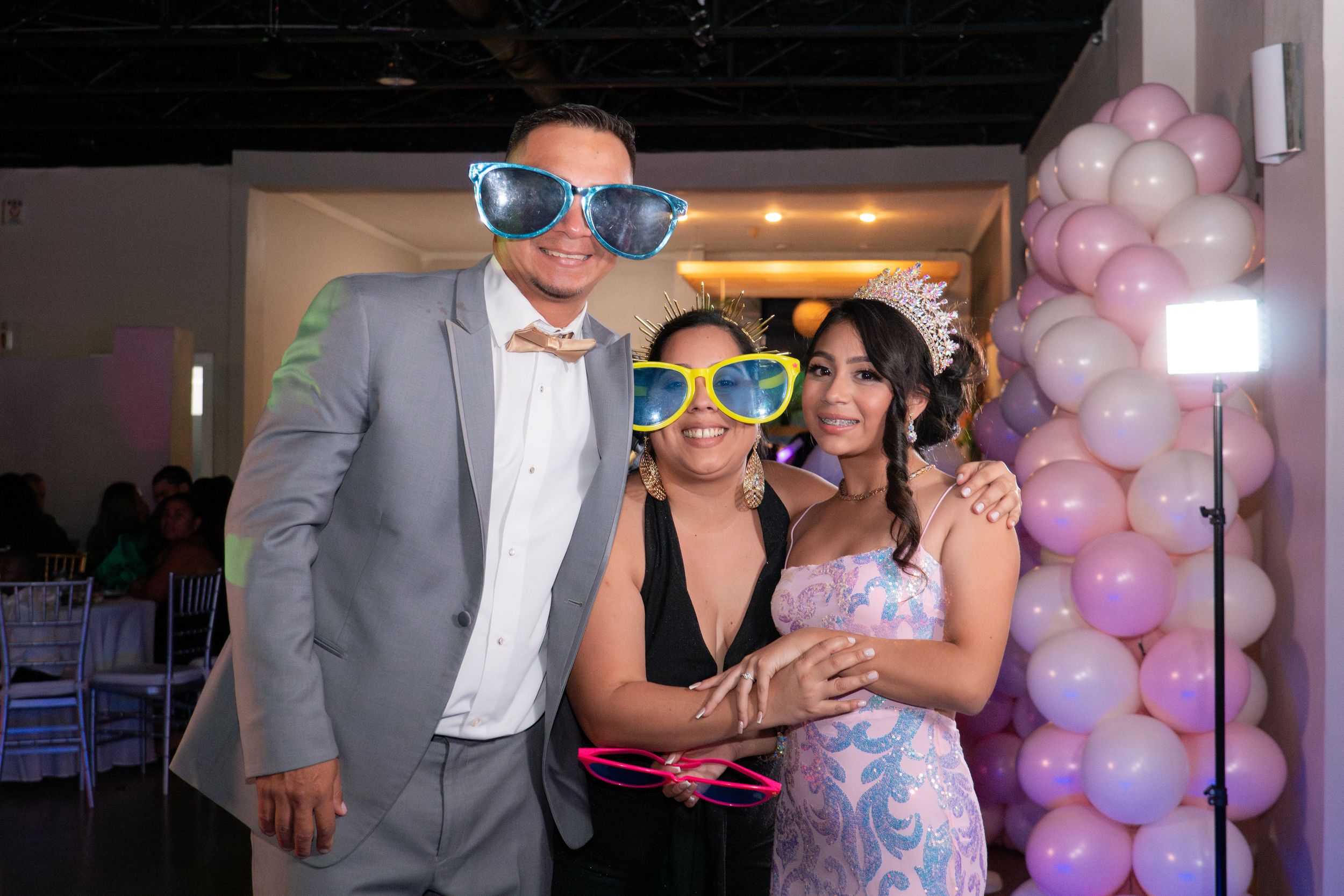 Three people pose with oversized novelty sunglasses at quinceañera celebration near pink and purple balloon column.