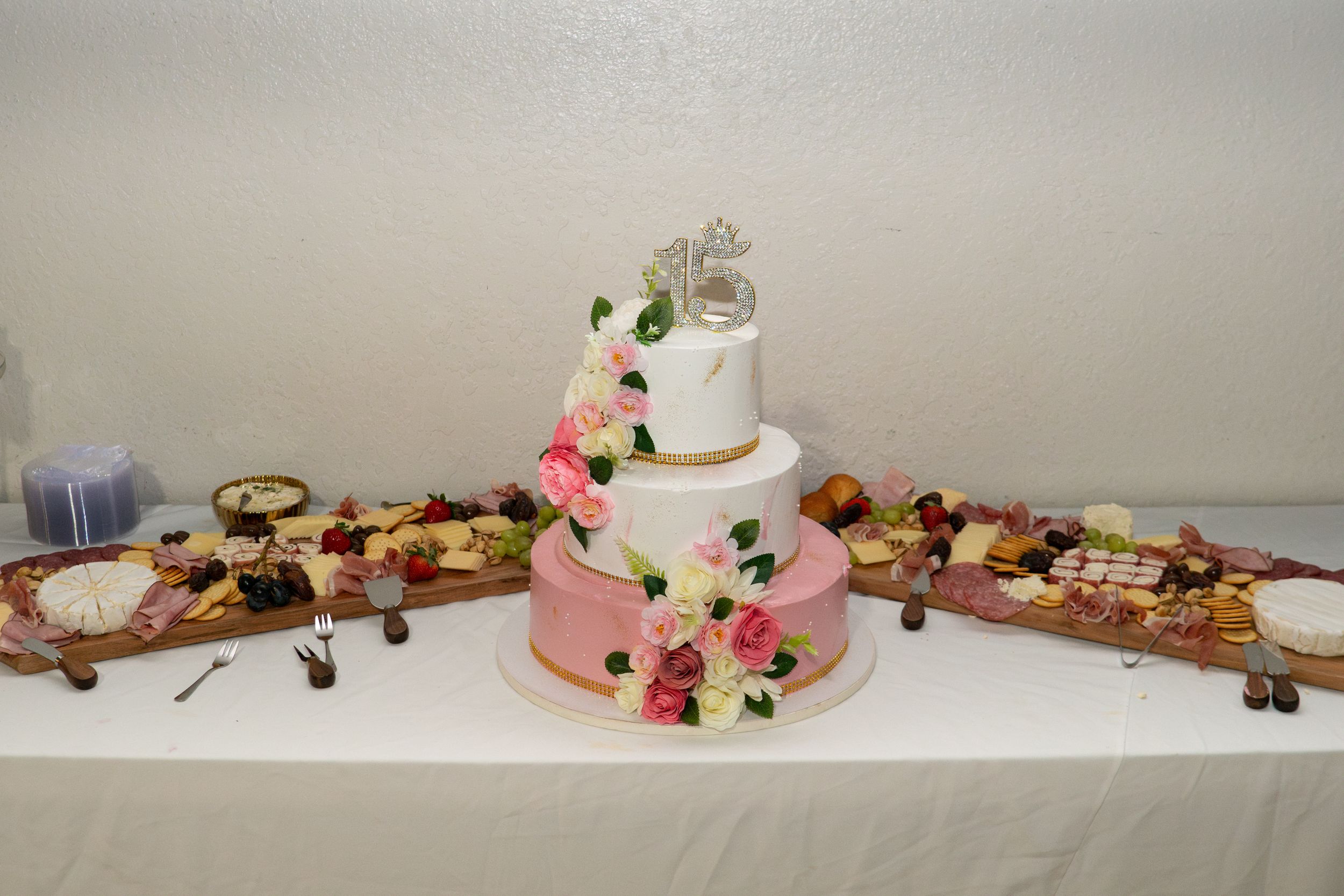 Three-tier quinceañera cake with pink roses and 15 topper surrounded by charcuterie boards on table.