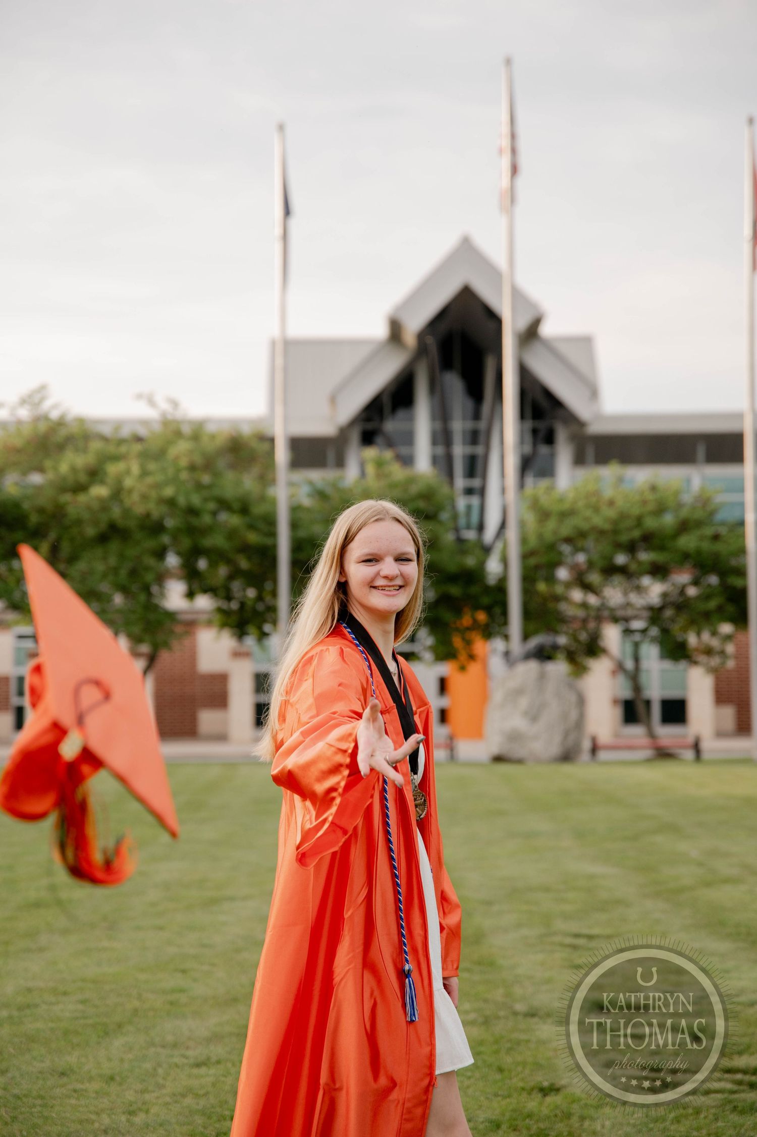 Graduate poses in bright orange graduation gown outdoors on campus lawn with buildings in background.