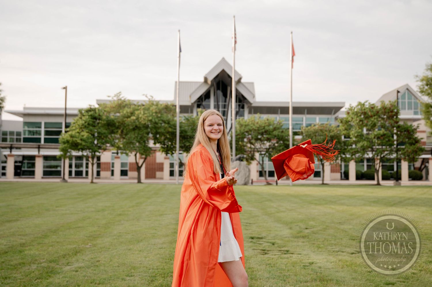 Someone in a bright orange graduation gown poses on a green lawn in front of a modern campus building.