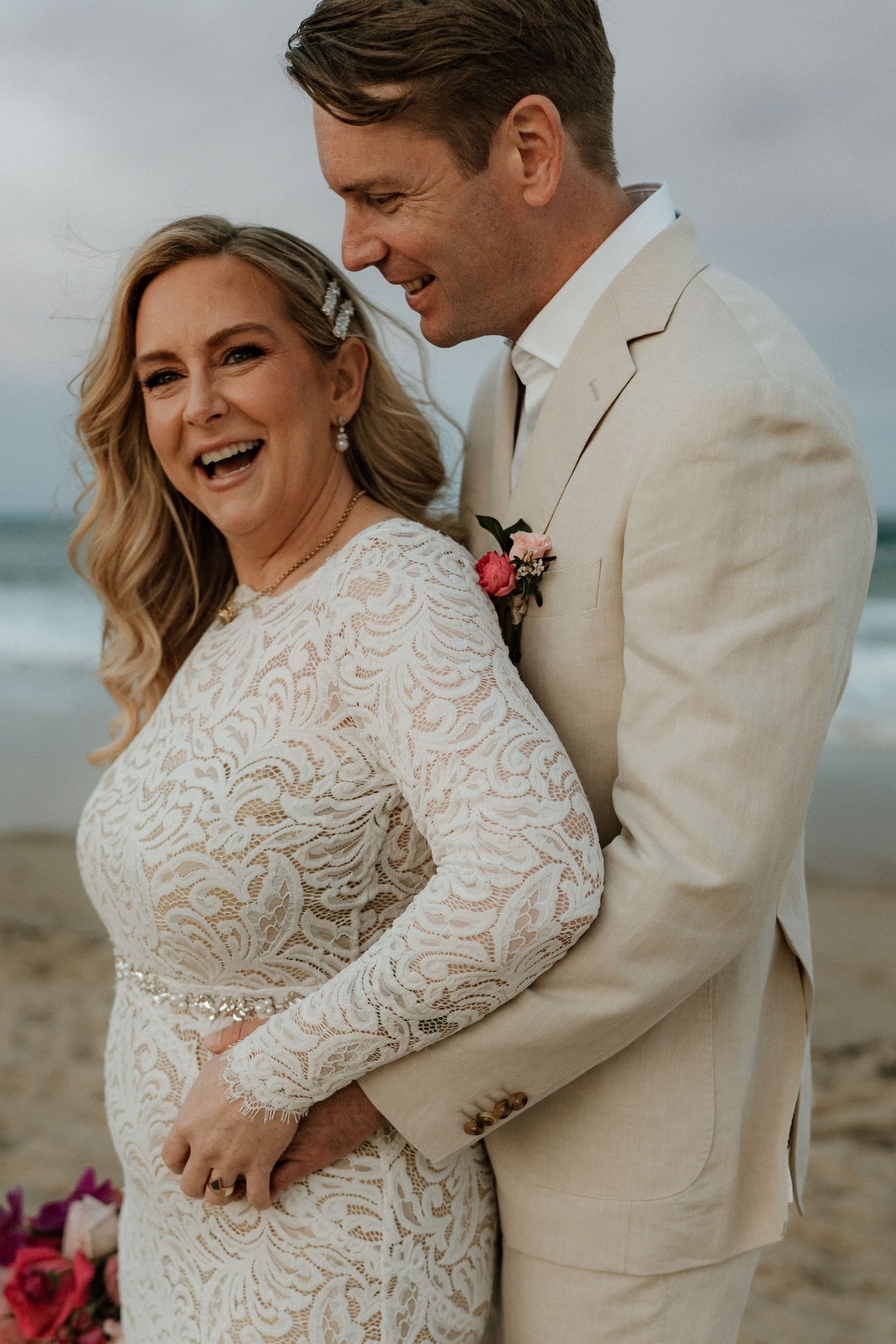 Bride in lace gown and groom in tan suit share a joyful moment on a beach wedding day.