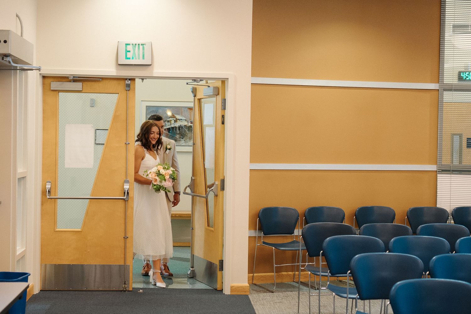 A beige waiting room with blue chairs and an exit sign mounted on the wall has medical equipment along the perimeter.