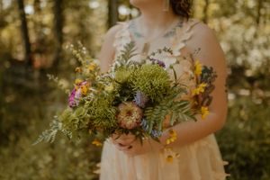 Bride in a sleeveless pink dress holding a bouquet of flowers in a forested area.