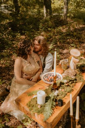 The couple embraces while seated on the forest floor beside a rustic wooden table set with candles and foliage.