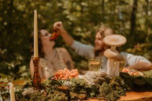 The couple, blurred in the background, shares grapes at a rustic table adorned with moss, mushrooms, and candles.