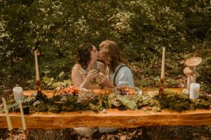 The couple shares a kiss at a rustic outdoor table adorned with greenery, candles, and an assortment of food.