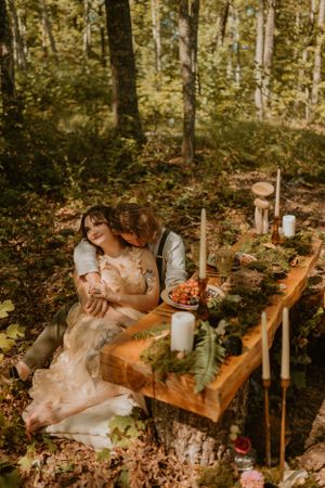 Newlyweds in the forest, groom hugging bride at rustic table with candles, flowers, and food after their elopement.