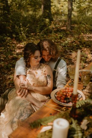 The couple sits embraced at their rustic reception table adorned with grapes and candles in a forest setting.