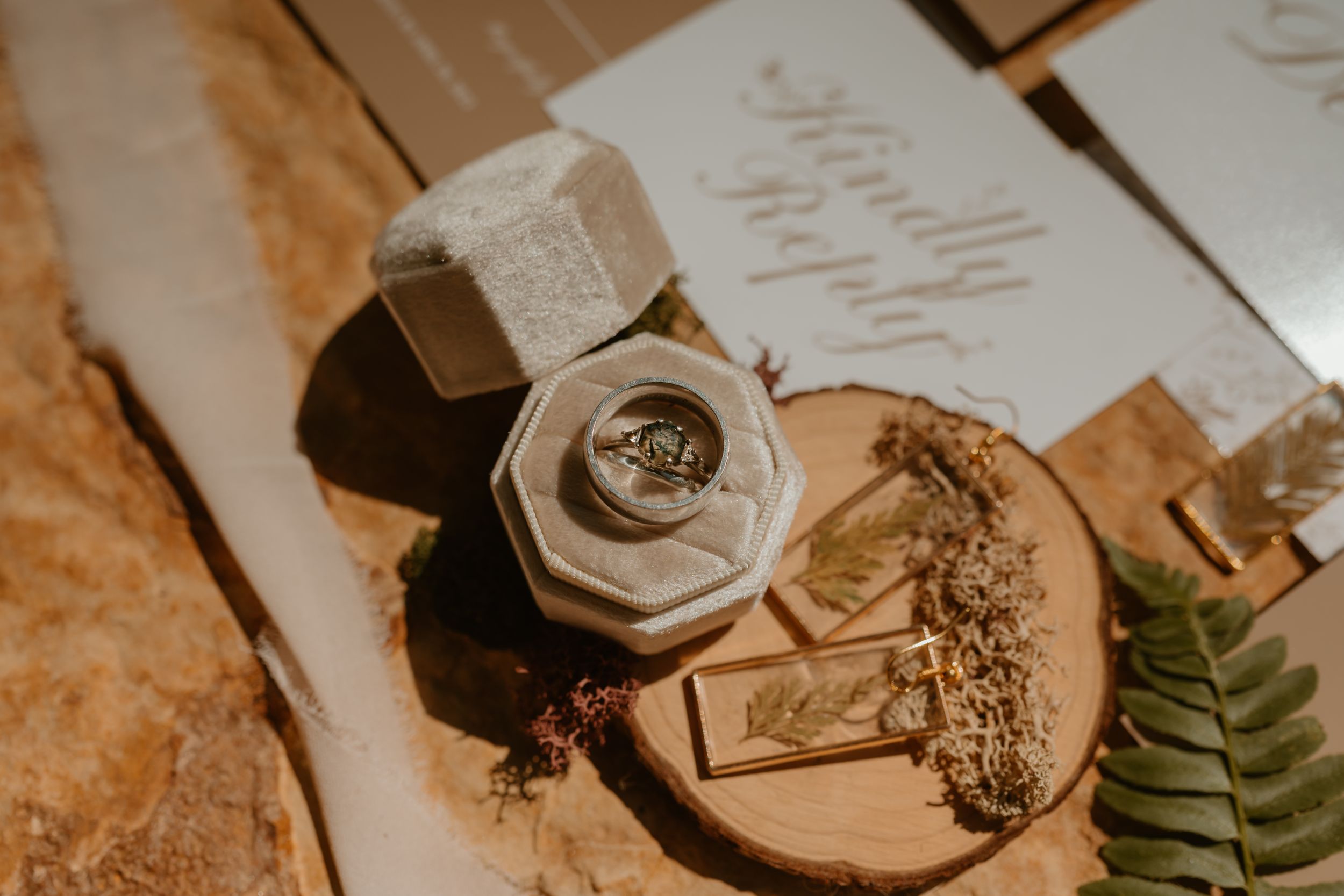 A velvet ring box with a silver ring on a wooden surface, next to dried flowers and a wedding invitation..