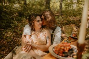 The couple sits closely, embracing each other affectionately in a forest setting with a picnic spread.