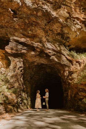 Kendall and Lane stand holding hands at the entrance of a rocky cave, surrounded by sunlight and shadows.