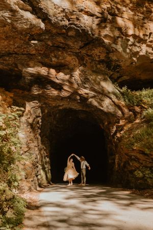The couple dances at the entrance of a large rocky cave, surrounded by greenery and sunlight.