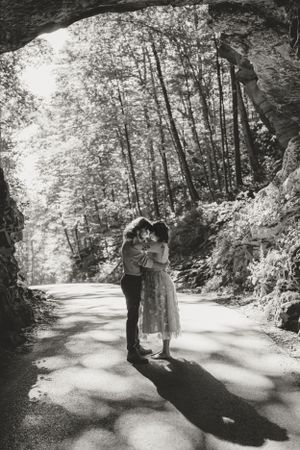 The couple embraces in the middle of a tree-lined path, sunlight casting shadows on the ground.