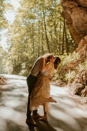 Lane kisses Kendall's neck on a sunlit forest road, with trees and rocks surrounding them.