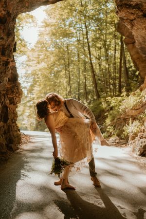 The bride and groom share a romantic dance dip beneath a sunlit tree canopy on a wooded path.