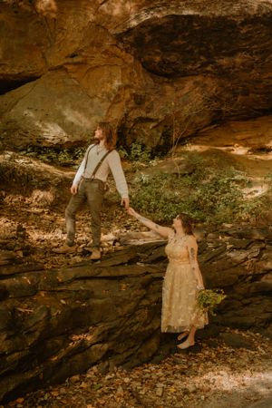 The couple stands on rocky terrain in a forest, holding hands. The bride holds a flower bouquet.
