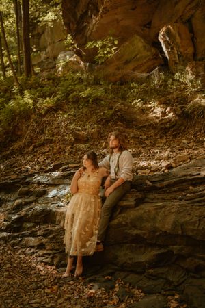 The bride and groom sit on rocks in a forest, with dappled sunlight filtering through the trees.