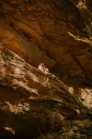 Bride and groom sit on a rocky ledge under a large cliff, surrounded by rugged natural rock formations and vegetation.