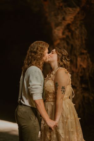 Bride and groom with long hair and rustic attire share a kiss while holding hands at their elopement ceremony..