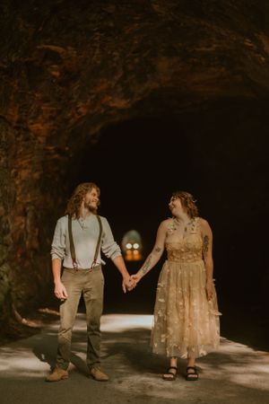 Bride and groom hold hands and smile at each other in a dimly lit tunnel, headlights entering from the opposite side.