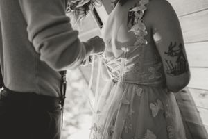 Close-up of groom tying the back of a lace-up wedding dress, with tattoos visible on the bride's arm.