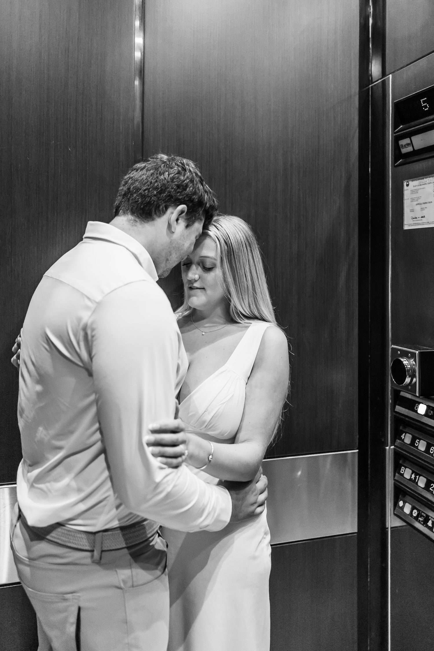 A couple in white wedding attire share romantic moments by an elevator door in a soft, warm lit hallway.
