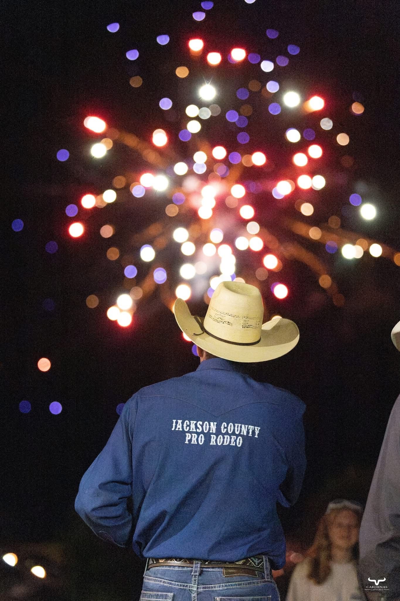 A group of people in blue uniforms and cowboy hats gather under colorful nighttime lights during an outdoor event.