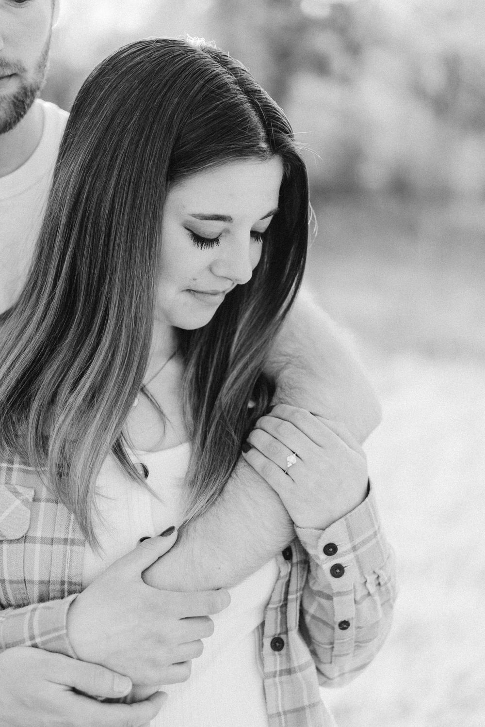 Black and white image of a quiet, intimate moment as she admires her ring, nestled into his arms, highlighting the emotion and stillness of