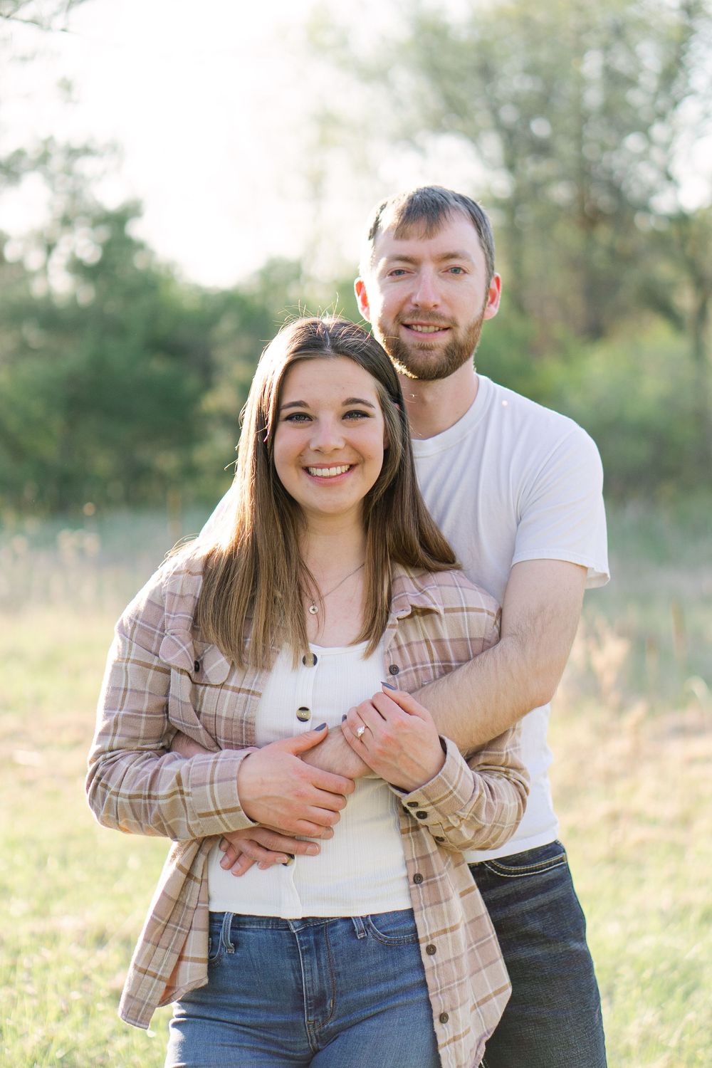 Country engagement session with a couple embracing in an open field, smiling warmly at the camera as golden light filters behind them