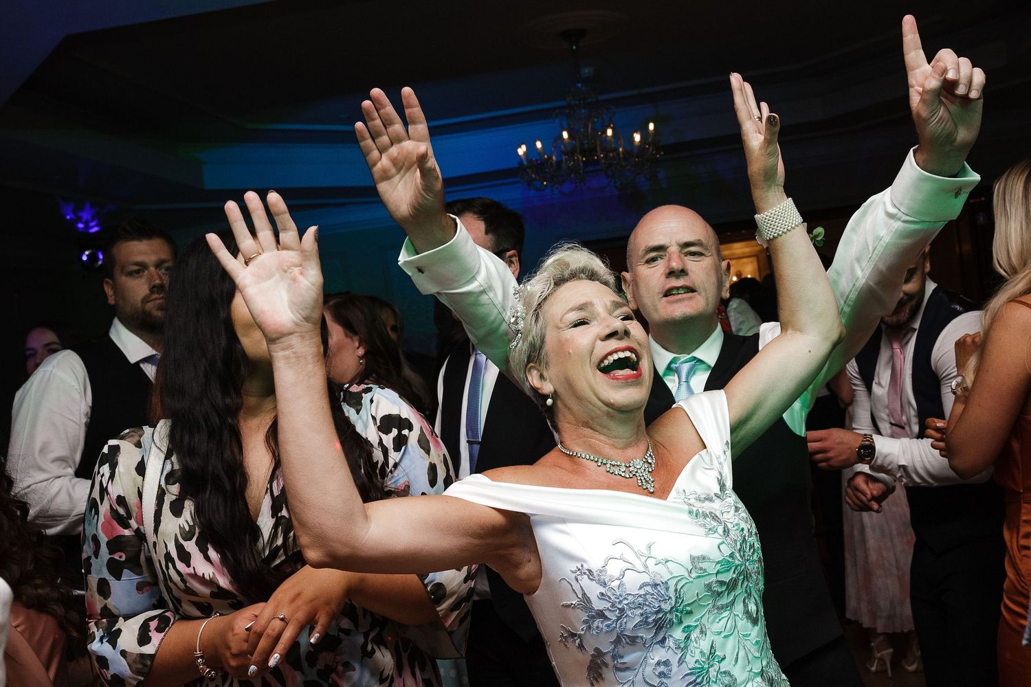 Energetic partygoers raise their hands and dance under blue lights at an event.