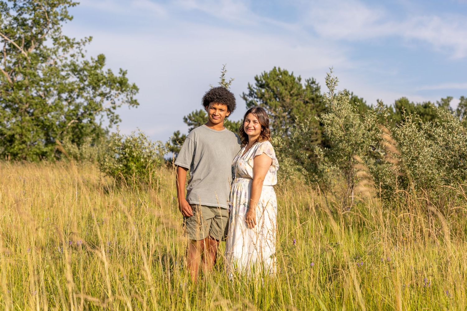 People walk through a sun-lit meadow with tall grass and scattered trees.