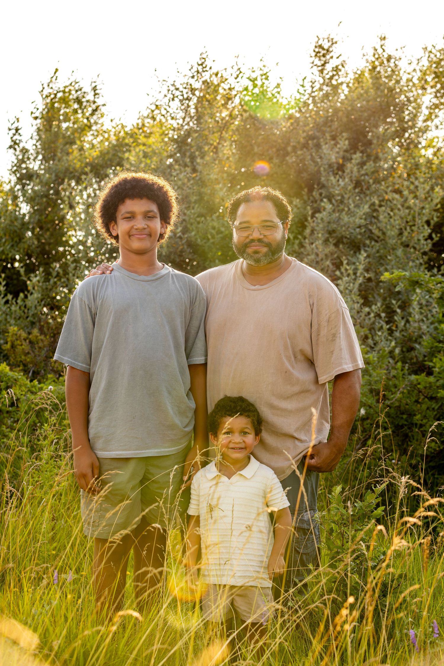 Three people standing together in a sunlit meadow with tall grass and trees.