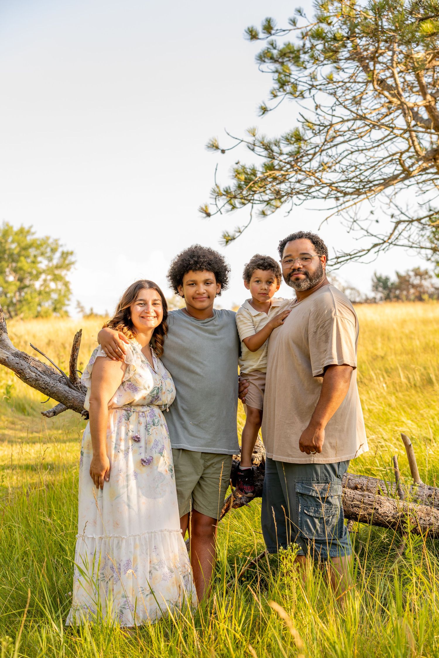 Family standing together by fallen tree in sunlit meadow with pine trees.