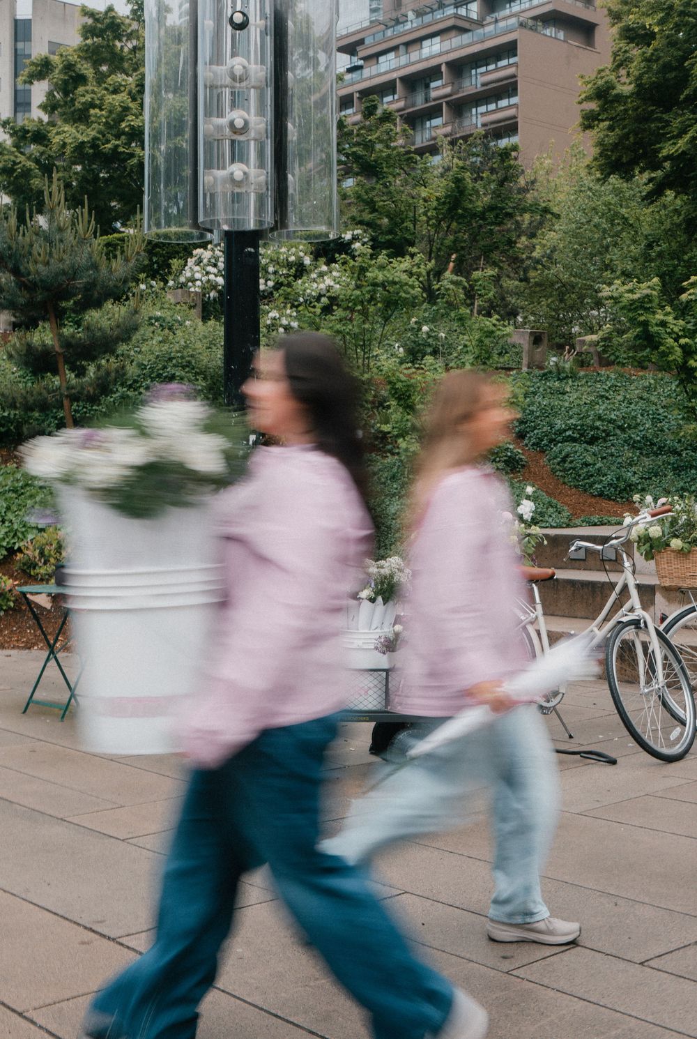 Motion blur of two people in pink sweaters walking past outdoor seating area with greenery and a parked bicycle.