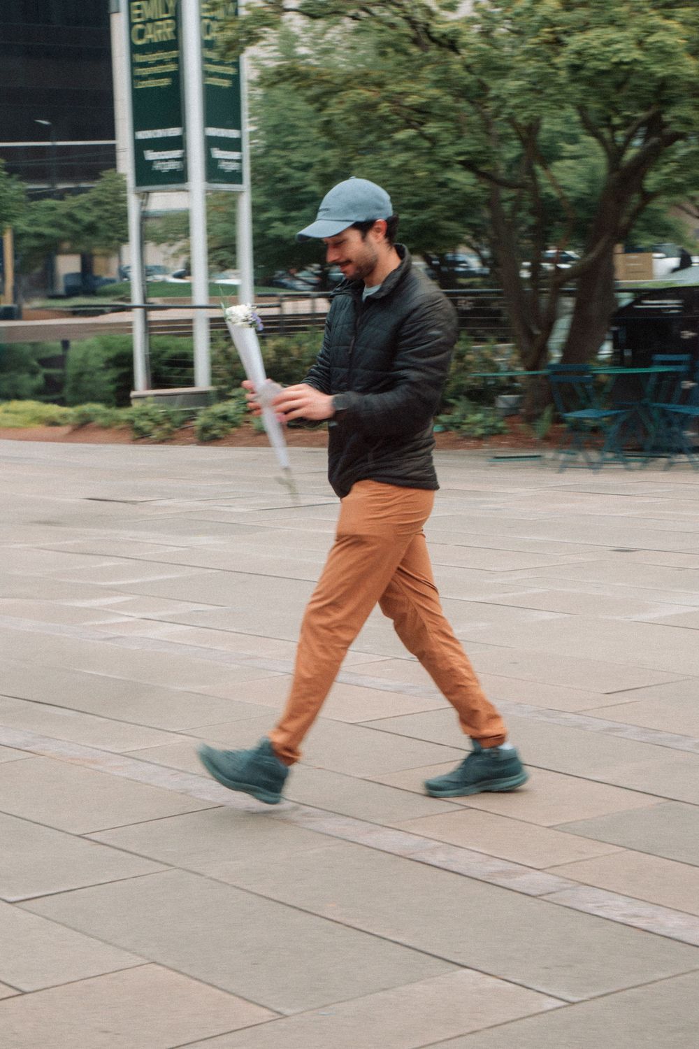 A person in orange pants and dark jacket walks across a tiled plaza while checking their phone.