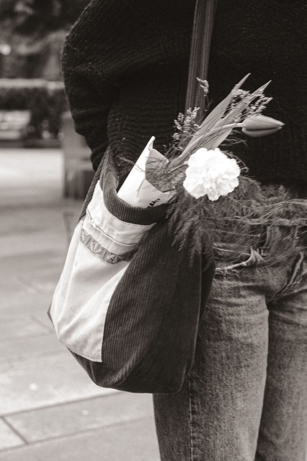 Black and white photo of a paper shopping bag with fresh flowers and baguette sticking out, carried against jeans.