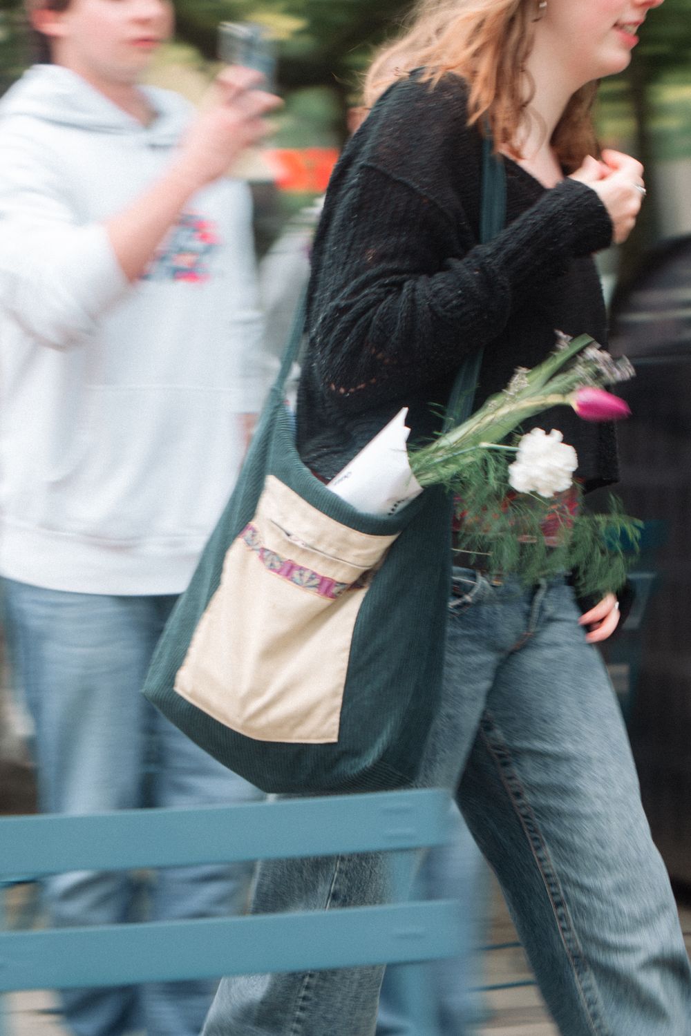 Person wearing black sweater and jeans carries canvas tote bag with fresh vegetables while walking outdoors.
