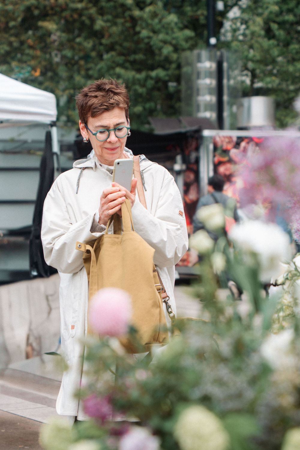 Person in glasses and white coat checks phone while holding brown tote bag outdoors with flowers in foreground.