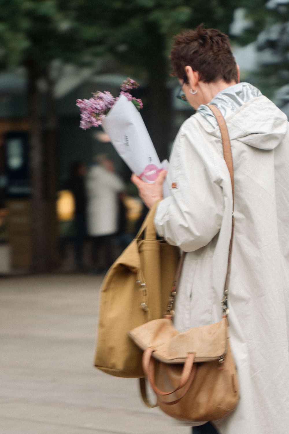 Someone in a light gray hoodie carrying shopping bags and pink flowers while walking on a city street.