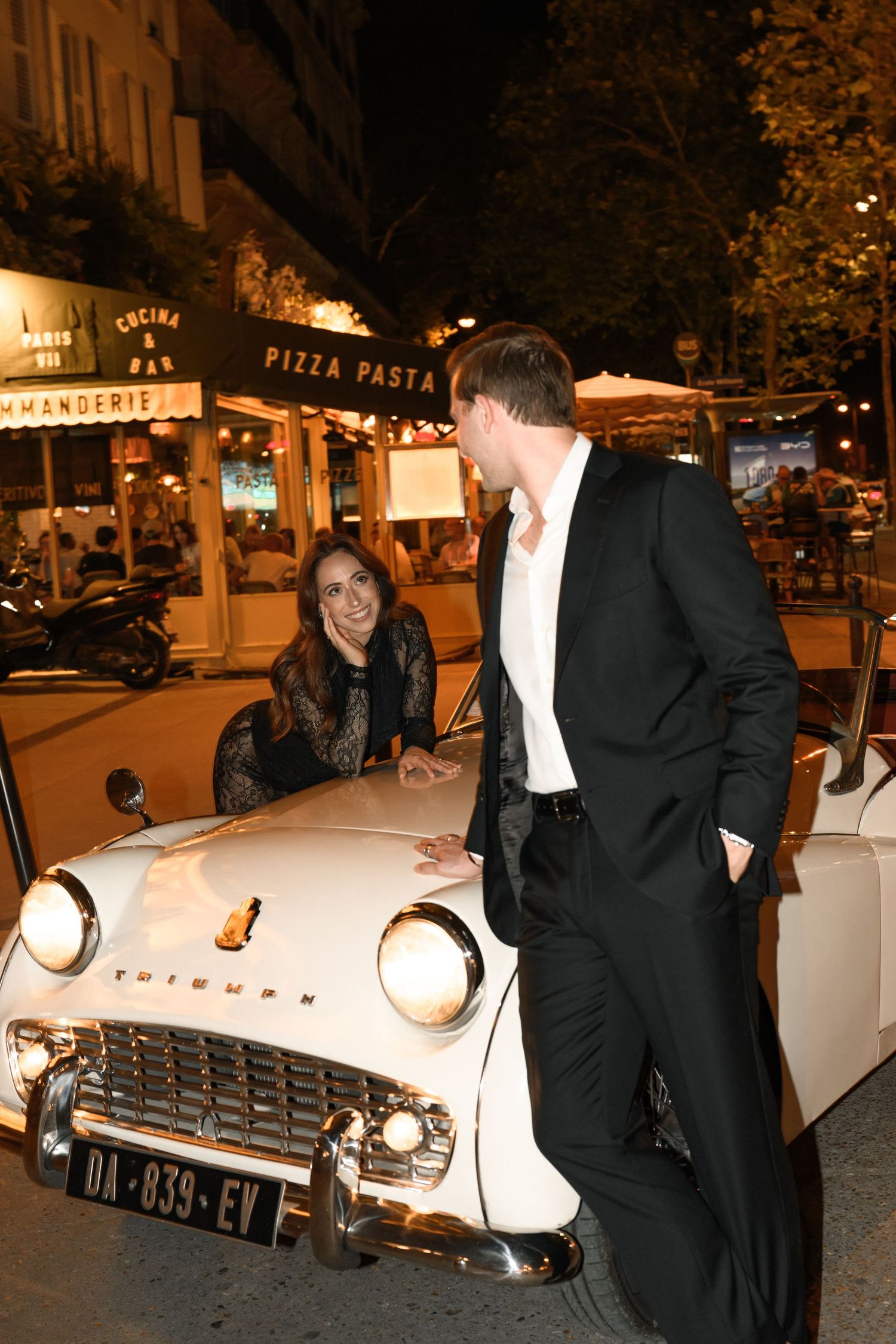A couple sits together in a classic red car wearing black outfits and sunglasses on a sunny day.
