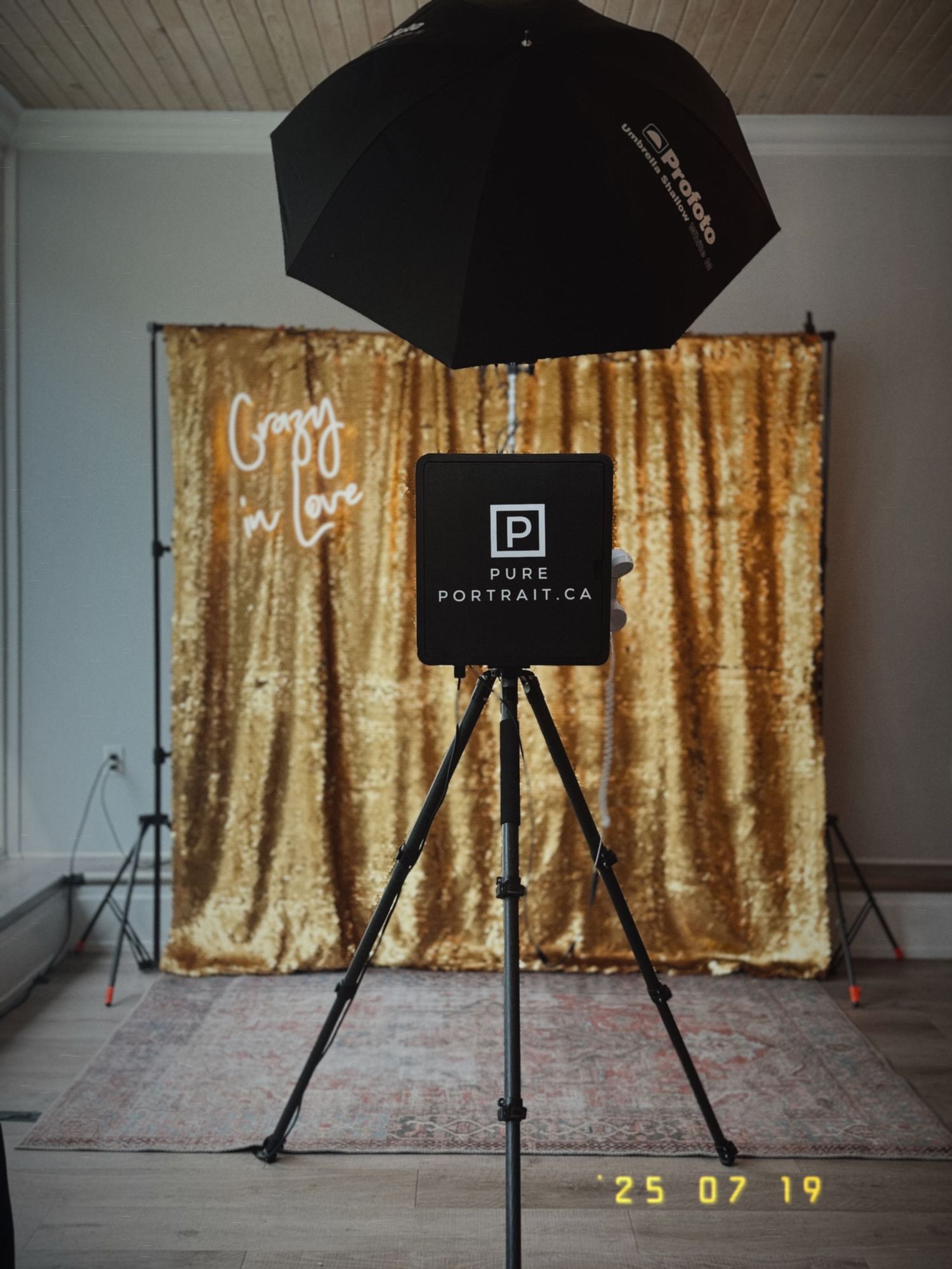 Photo booth setup with black umbrella light and gold sequin backdrop against a white wall with Portratica branding.