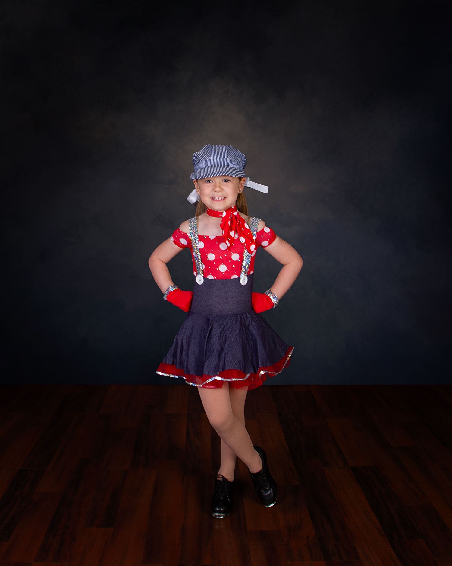 Young dancer in a pink tutu and black leotard poses gracefully on a gray ottoman against a dark backdrop.
