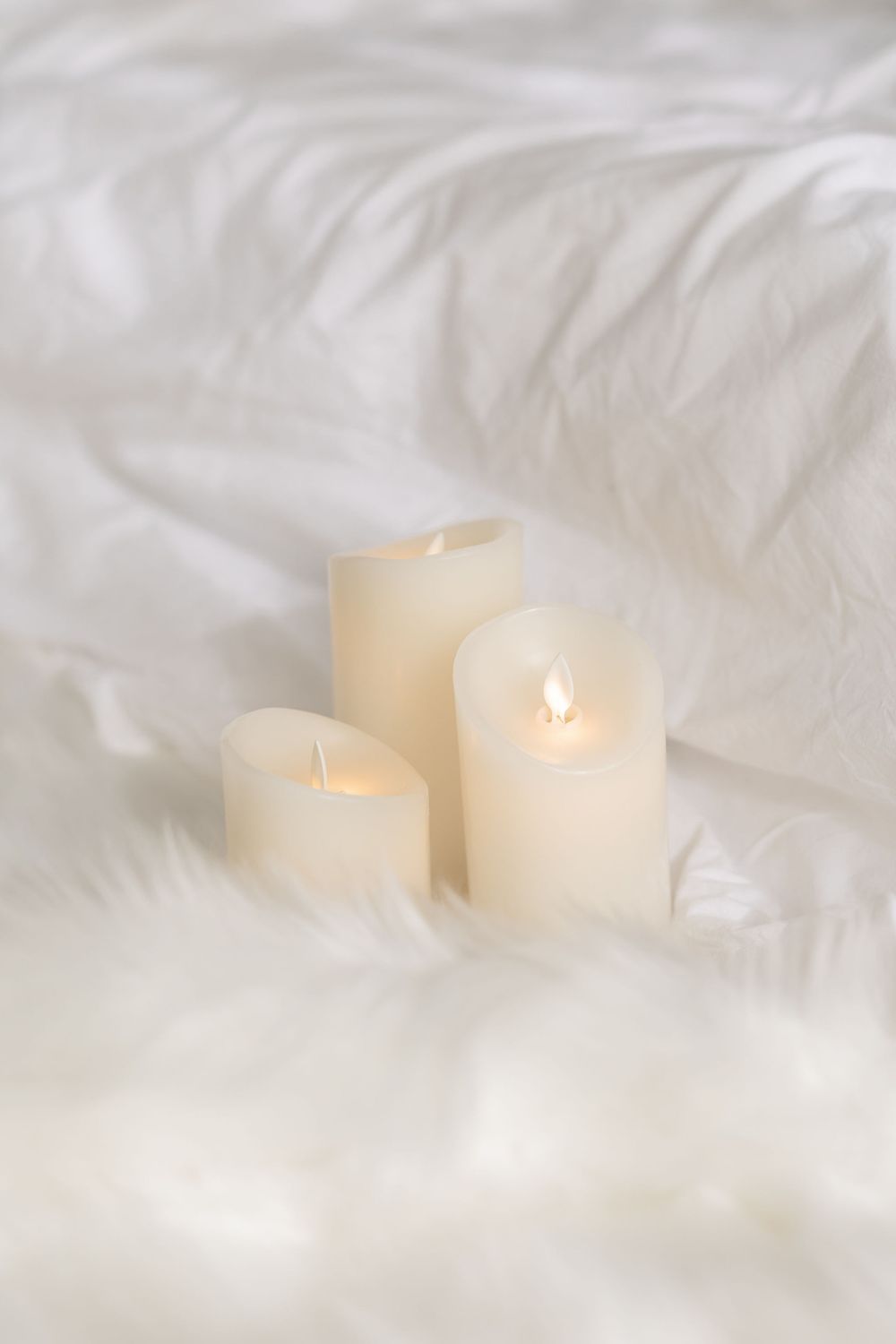 Three white pillar candles glowing softly against a plush white fabric background.