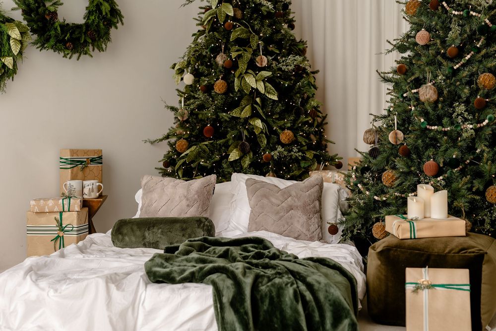 Cozy bedroom decorated for Christmas with multiple trees, white bedding, green throw blanket and wrapped presents.