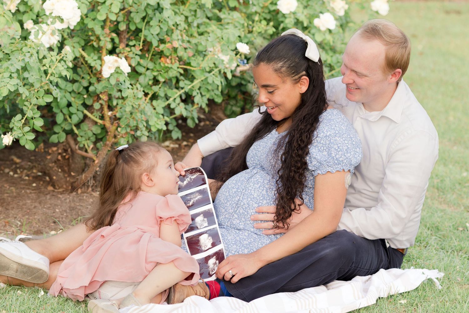 Family of 3 sitting down in the grass with dad behind mom and toddler daughter in front lying on pregnant mom smiling at ultrasound photos.