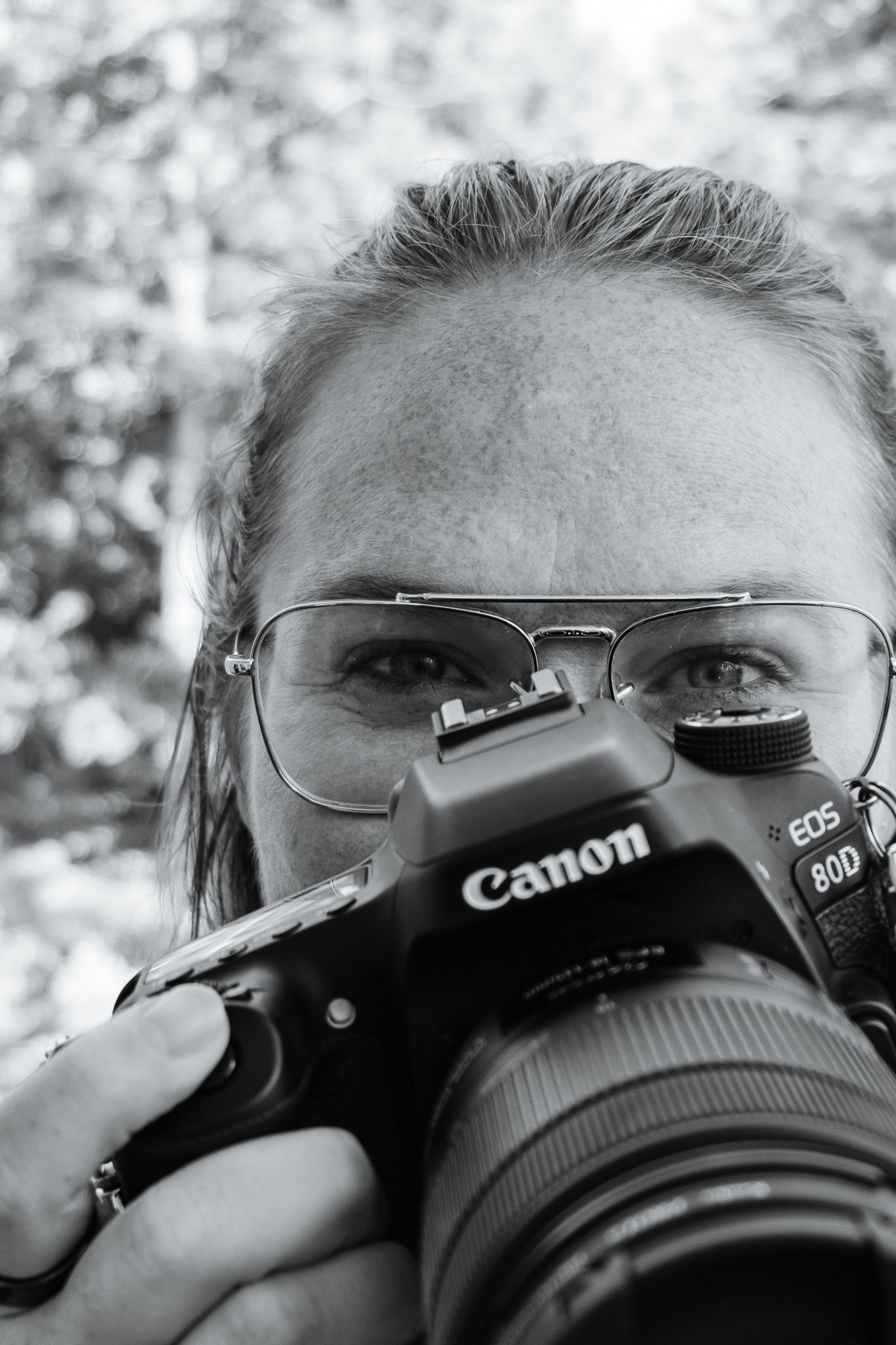 Black and white photo of a woman wearing glasses holding a canon camera up close to her face just under her eyes.