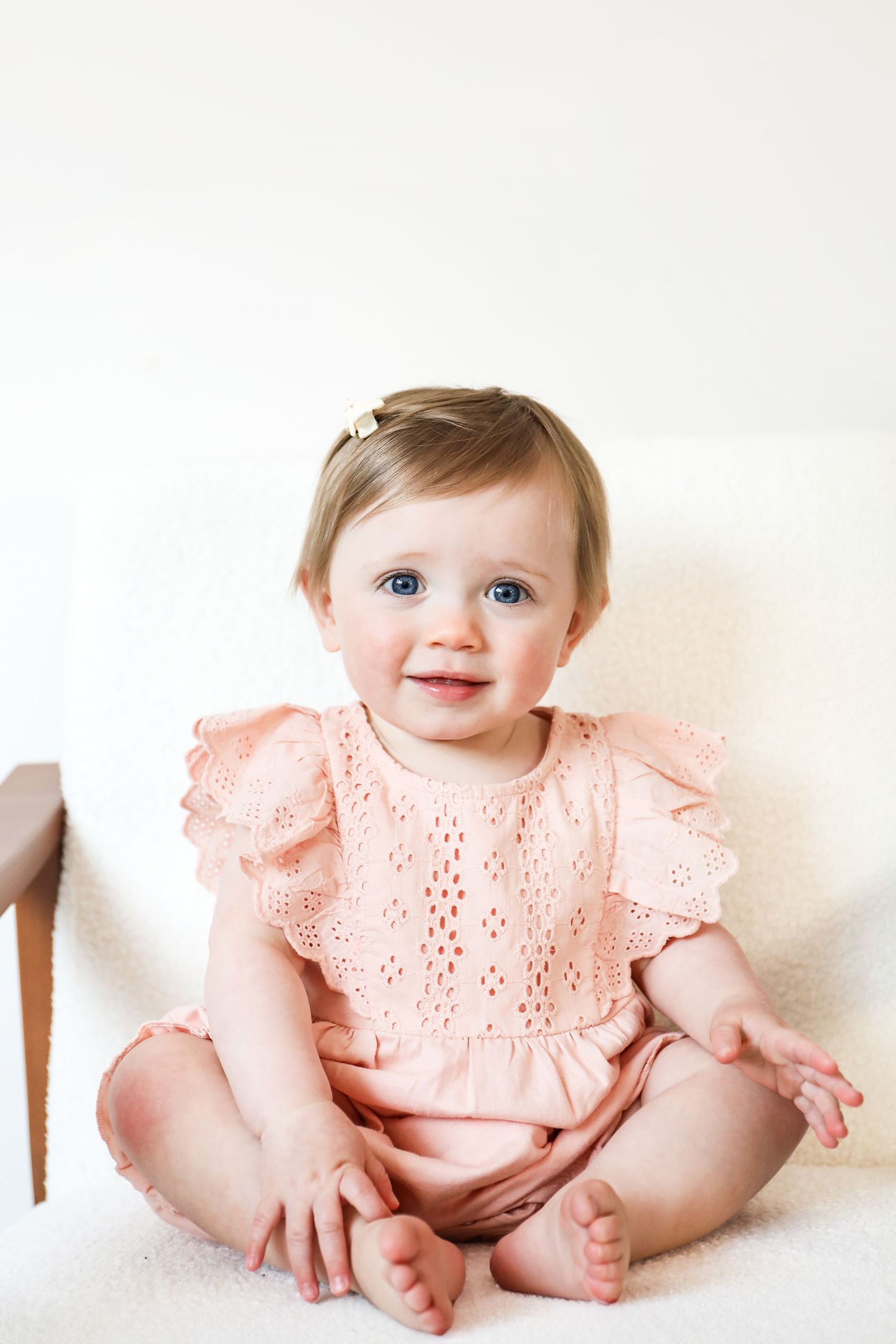 A baby wearing a peachy-pink dress sits on white seat during birthday shoot in Nottingham